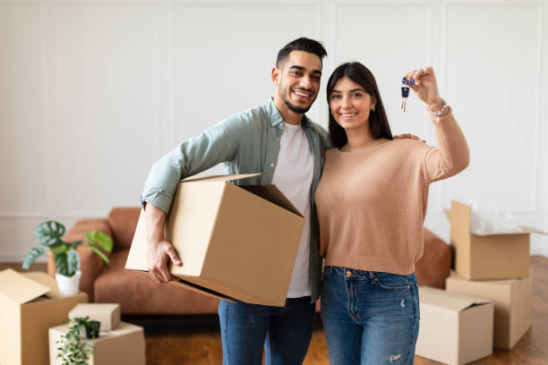 A smiling couple holding a cardboard box and keys, standing in a room with moving boxes.