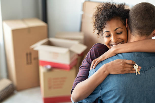 A couple embraces joyfully in front of moving boxes, one holding a set of keys.