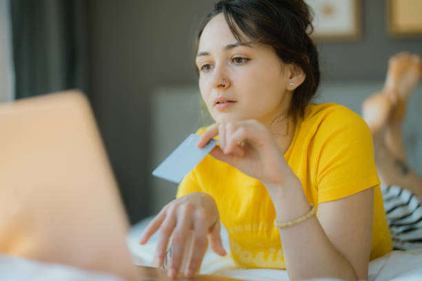 A woman in a yellow shirt lies on a bed while looking at a laptop and holding a credit card, researching apartments.